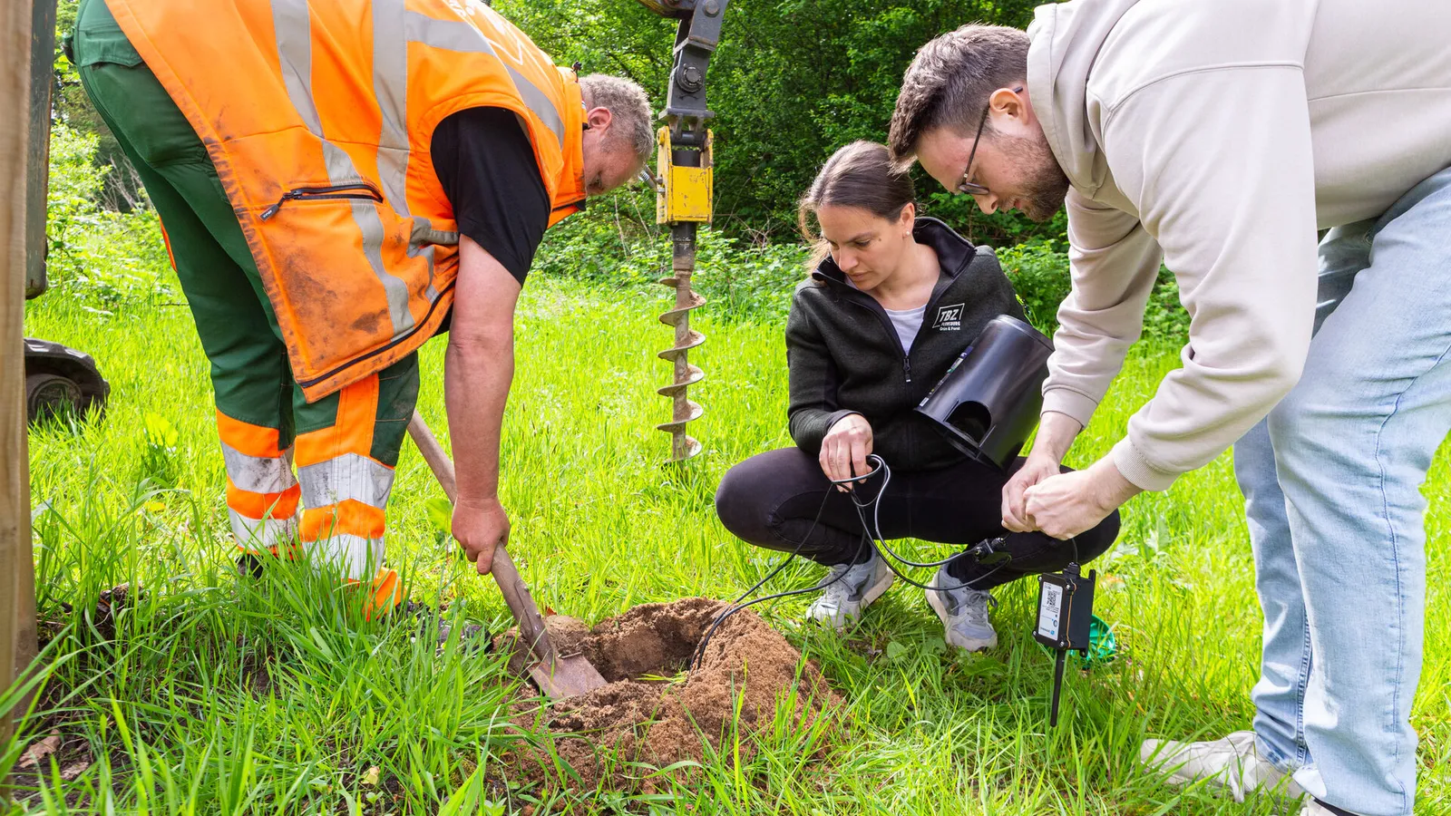 At a freshly dug hole one person backfills with a spade while two others connect the sensor unit, the earth auger hanging in the background