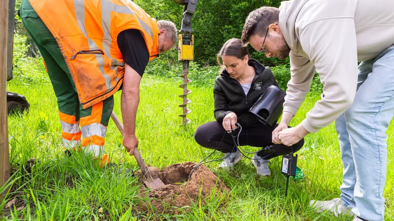 At a freshly dug hole one person backfills with a spade while two others connect the sensor unit, the earth auger hanging in the background