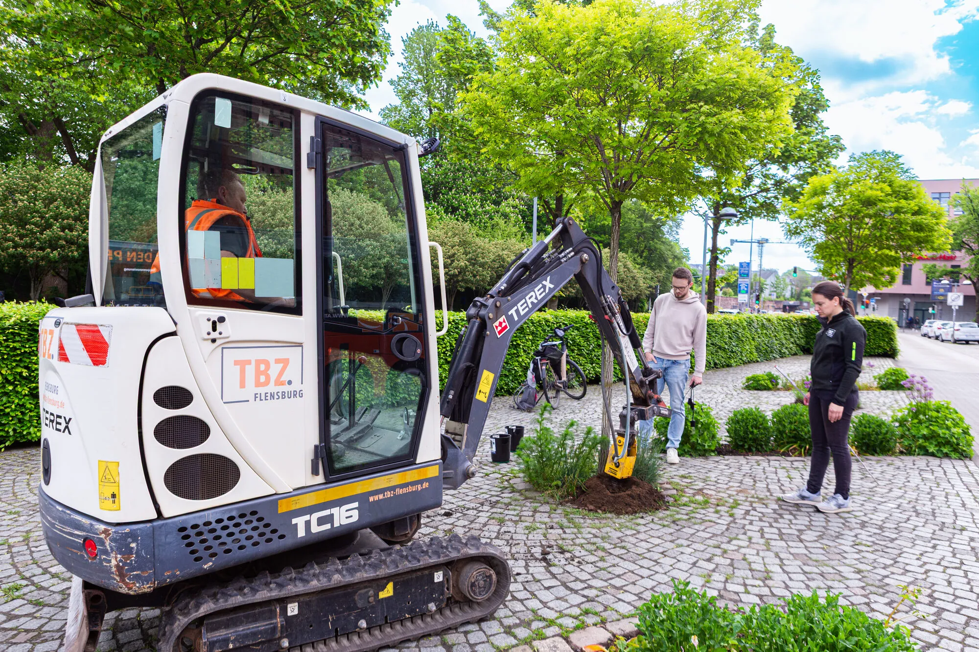 A mini excavator belonging to the Flensburg municipal operations centre at a tree pit on a street, with two people standing beside it.