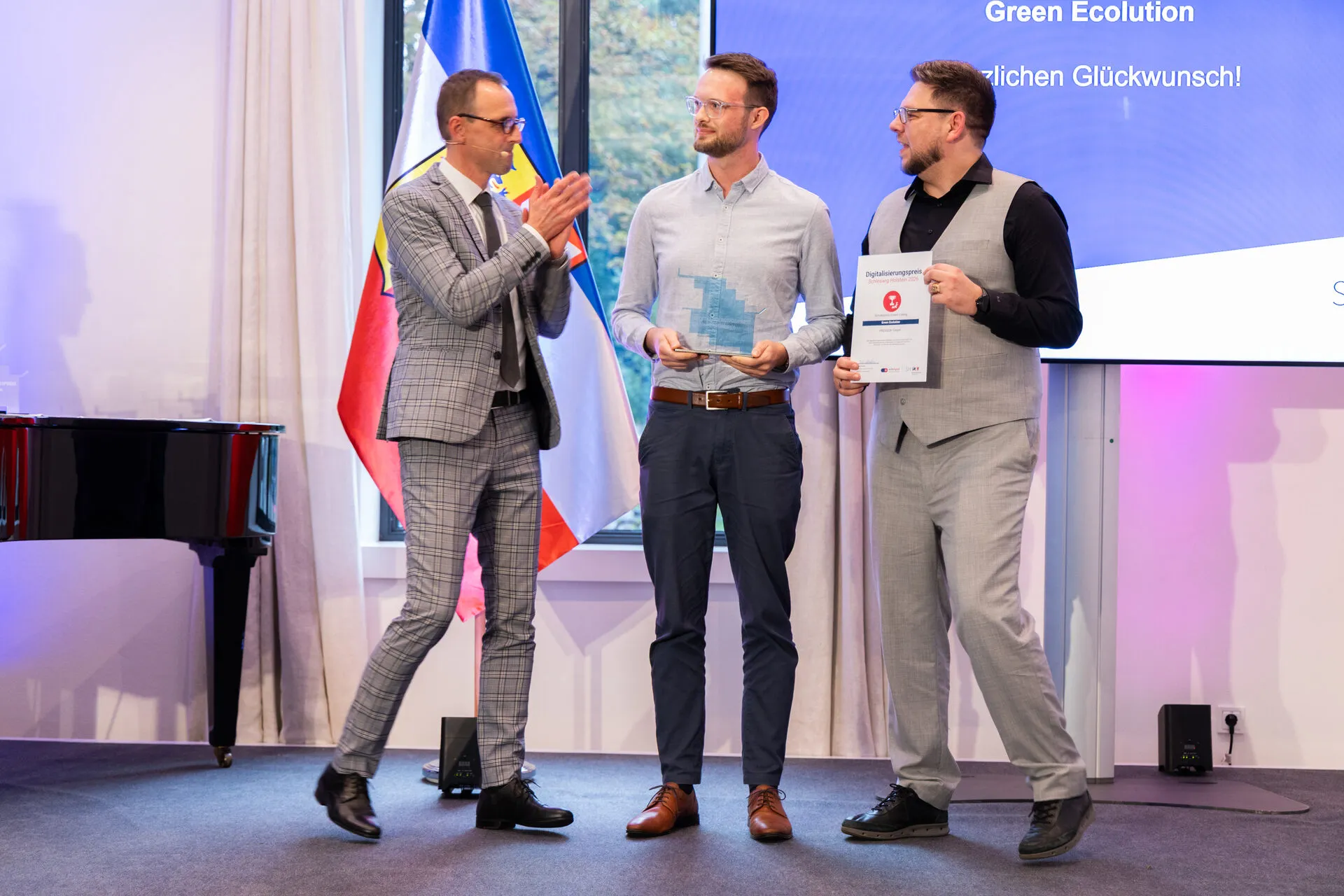 Digital minister Dirk Schrödter applauds two PROGEEK representatives holding trophy and certificate on stage, behind them a screen reading Green Ecolution