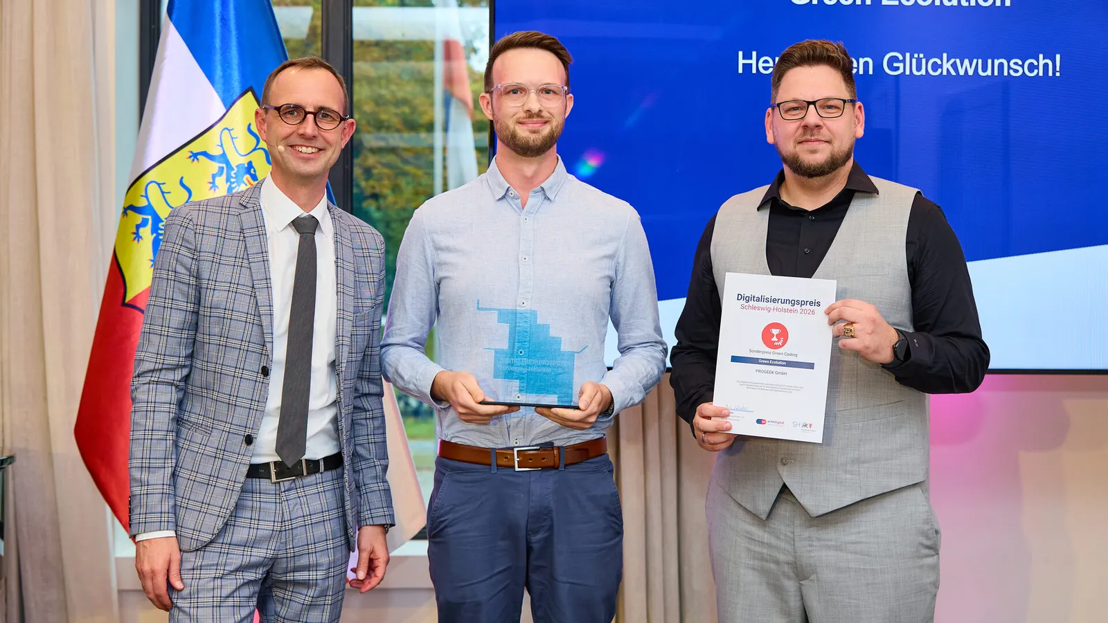 Digital minister Dirk Schrödter stands with two PROGEEK representatives in front of a screen reading "Green Ecolution, Herzlichen Glückwunsch!", one holds the glass trophy, the other the certificate