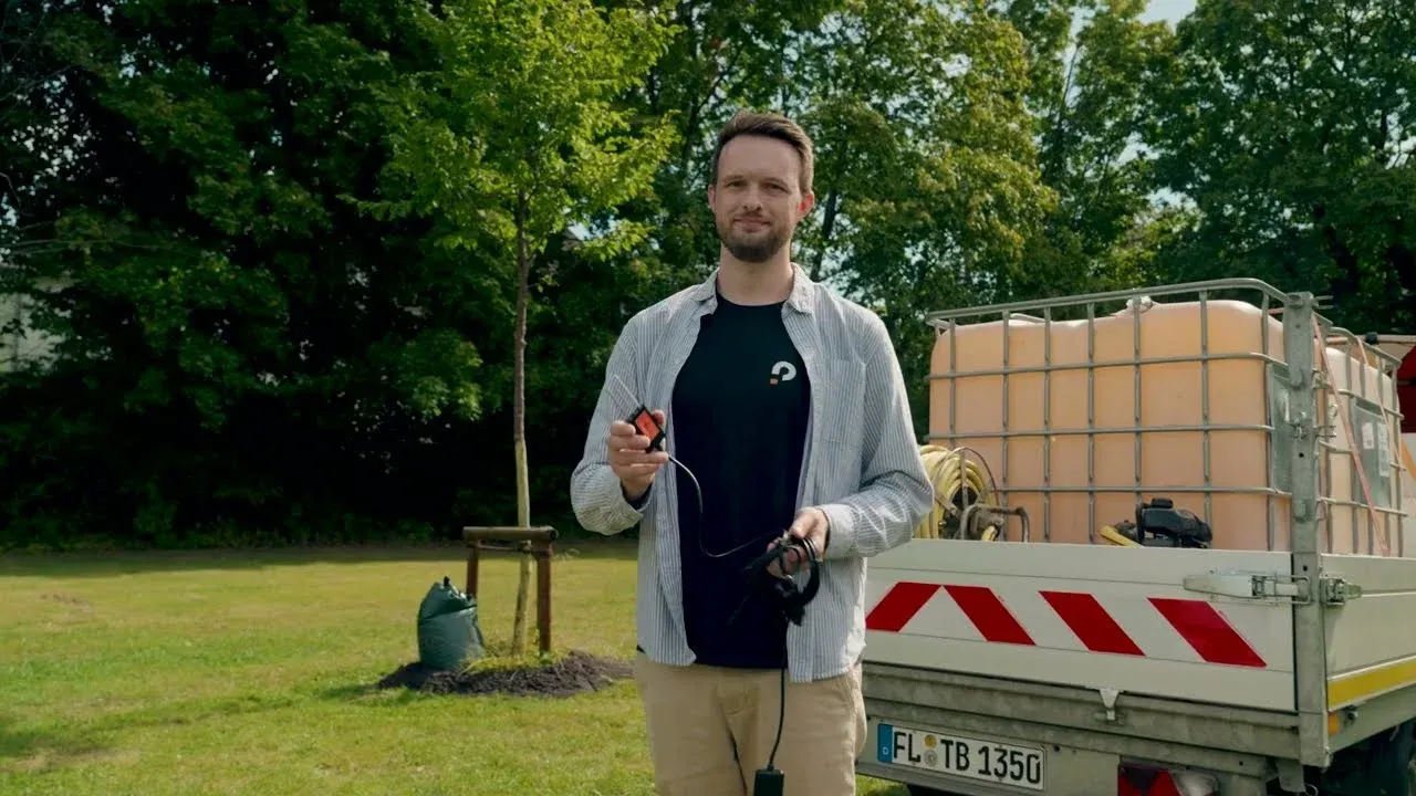 A project member holding a soil moisture sensor, with a young street tree and a flatbed truck with a water tank in the background