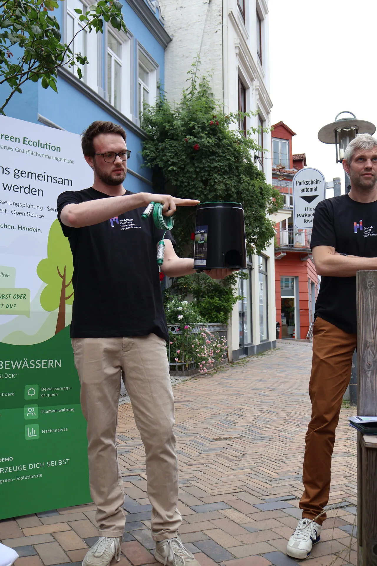 Cedrik Hoffmann holds up a ground enclosure box and a Watermark sensor in front of the Green Ecolution roll-up while explaining them, with Simon Olberding standing to his right
