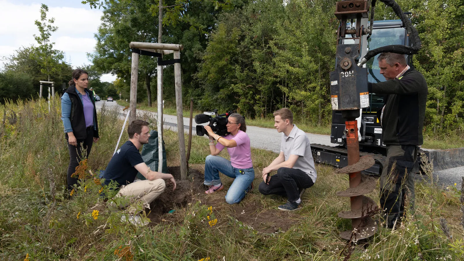 A camera team films the installation of a soil moisture sensor at the roadside, two people kneel beside the tree, and the earth auger of an excavator stands in the hole on the right